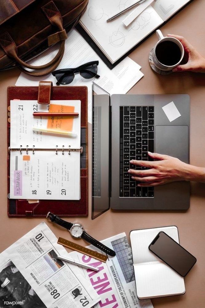 Overhead view of a desk with a laptop, an open planner, a newspaper, a phone, a coffee cup and sunglasses.